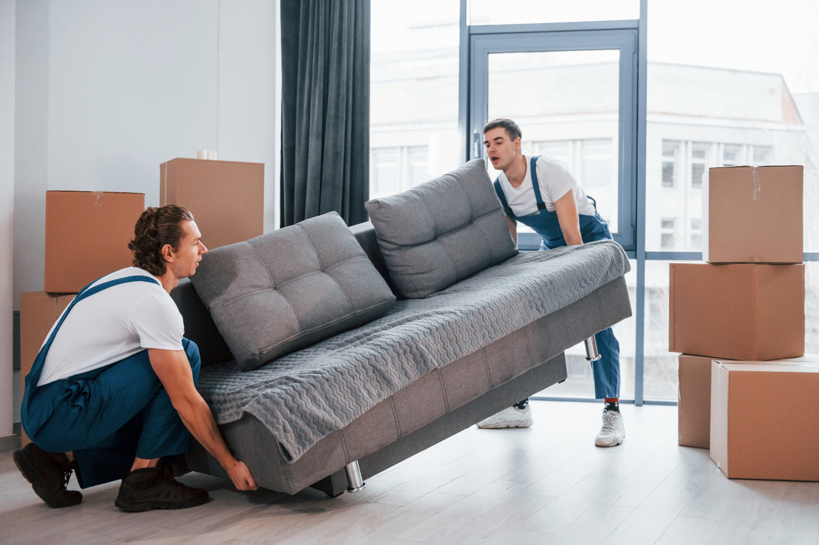 Wood Floor Sanding in south-west-london — two movers lift a grey sofa back onto newly refinished light oak engineered plank flooring, with packing boxes in a bright room.